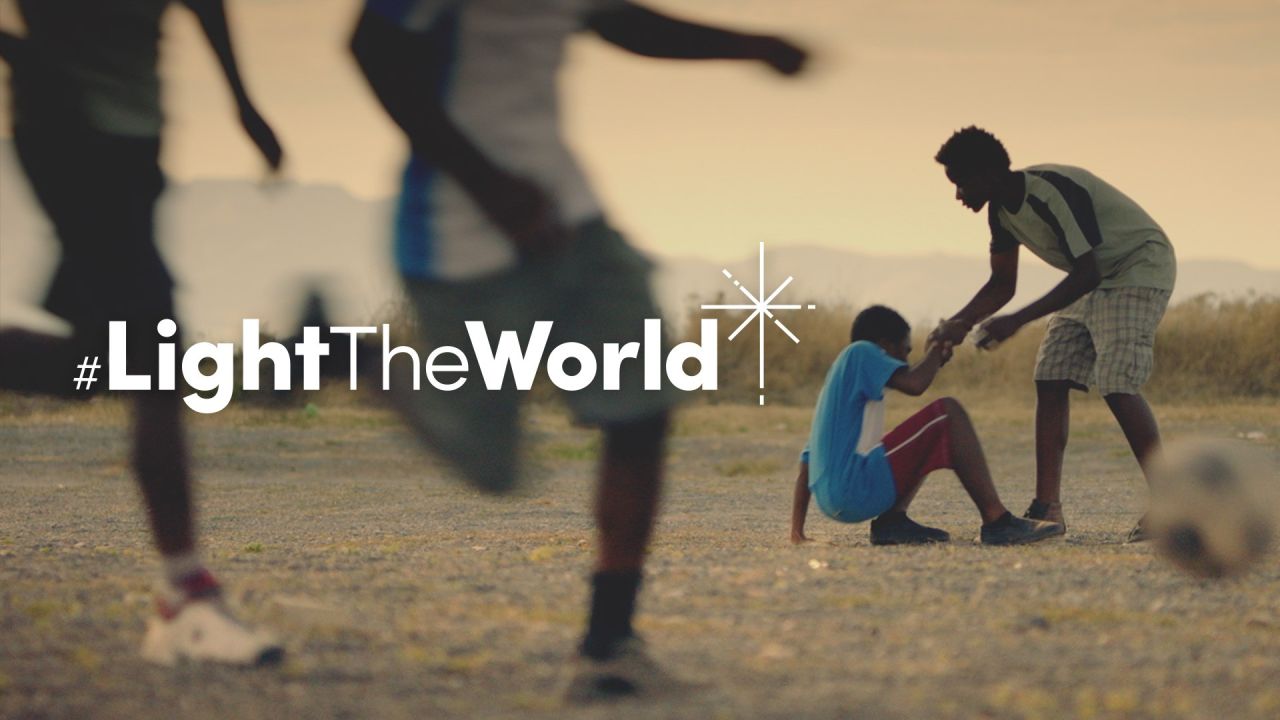 A boy helps another boy get up after having fallen over playing soccer from the Light the World video